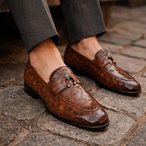 May include: Brown leather loafers with a woven pattern and a decorative buckle. The shoes are worn with gray trousers, and are sitting on a cobblestone surface. The loafers have a glossy finish.