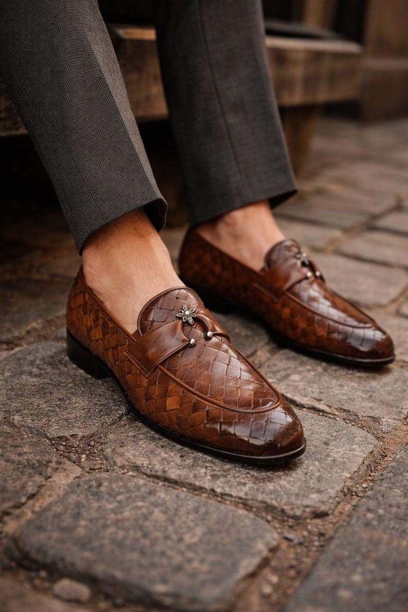 May include: Brown leather loafers with a woven pattern and a decorative buckle. The shoes are worn with gray trousers, and are sitting on a cobblestone surface. The loafers have a glossy finish.