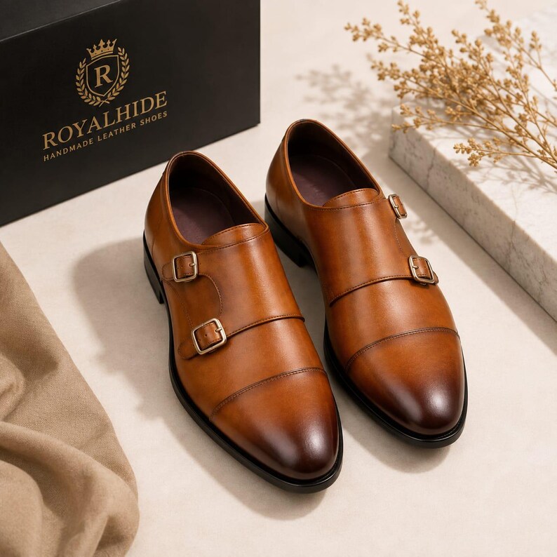 May include: A pair of tan leather double monk strap shoes with a cap toe design and gold-toned buckles. The shoes are positioned near a black box featuring the gold text ROYALHIDE HANDMADE LEATHER SHOES and a crown logo with the letter R.