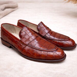 May include: A pair of brown leather loafers with a textured, crocodile-like pattern. The shoes have a classic design with a rounded toe and a low heel. The loafers are displayed on a neutral surface.
