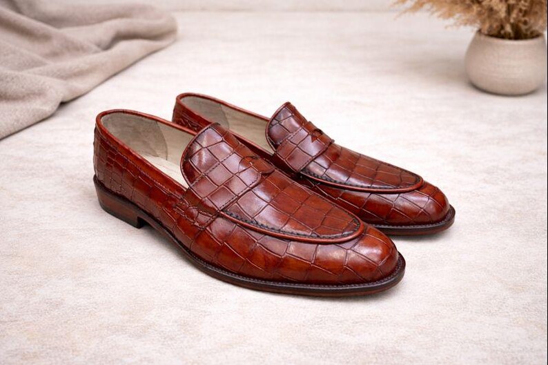 May include: A pair of brown leather loafers with a textured, crocodile-like pattern. The shoes have a classic design with a rounded toe and a low heel. The loafers are displayed on a neutral surface.
