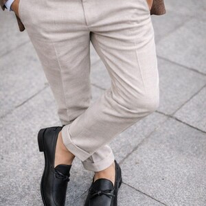 May include: A person wearing a brown blazer, light blue shirt, and beige pants. The person is wearing a gold watch and black leather loafers with a bow detail. The pants are cuffed at the ankles.
