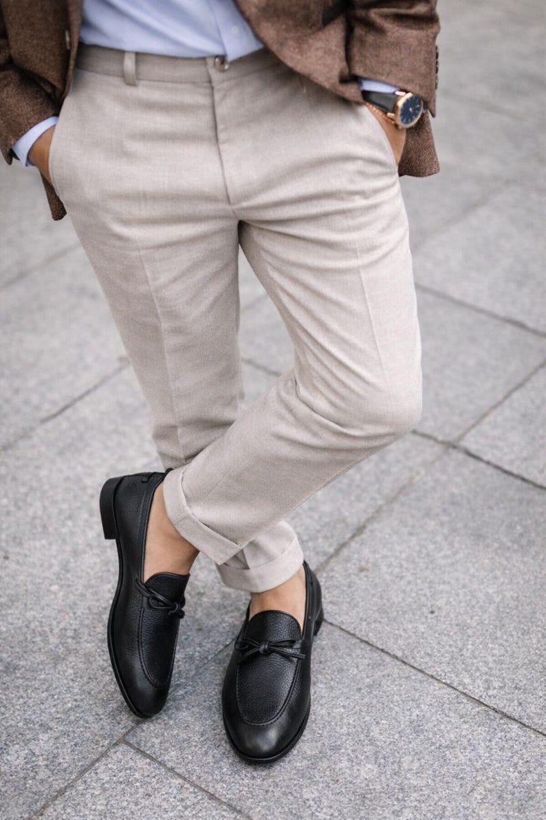 May include: A person wearing a brown blazer, light blue shirt, and beige pants. The person is wearing a gold watch and black leather loafers with a bow detail. The pants are cuffed at the ankles.