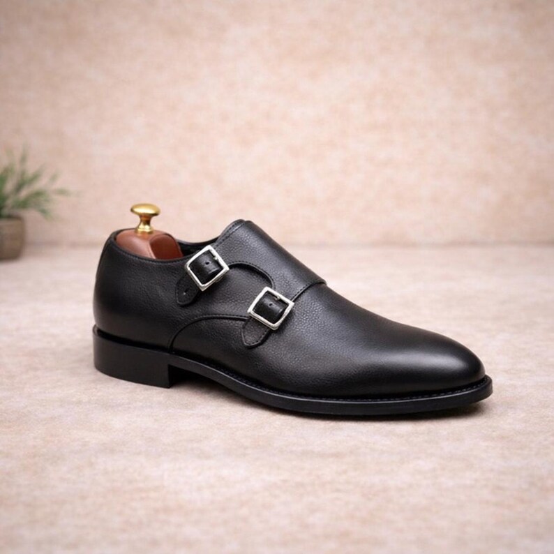May include: Black leather double monk strap shoe with silver buckles. The shoe has a rounded toe and a low heel. The shoe is displayed on a neutral background.