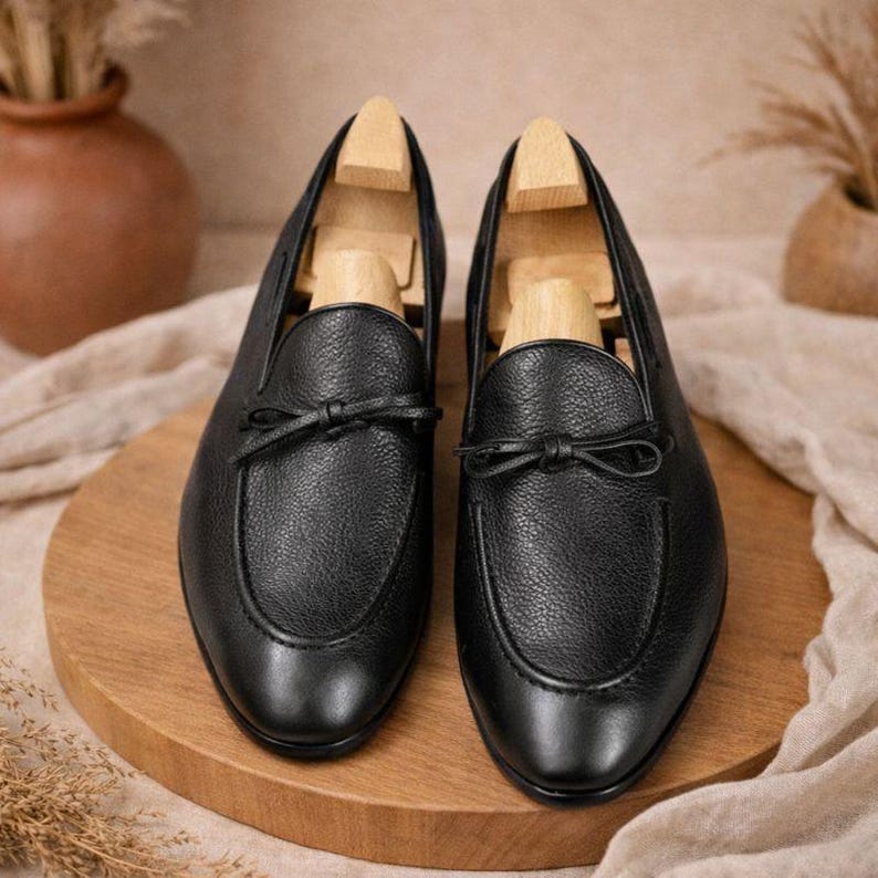 May include: A pair of black leather loafers with a textured finish. Each shoe features a small bow detail on the vamp and is displayed with a wooden shoe tree inside. The shoes are set on a wooden surface.
