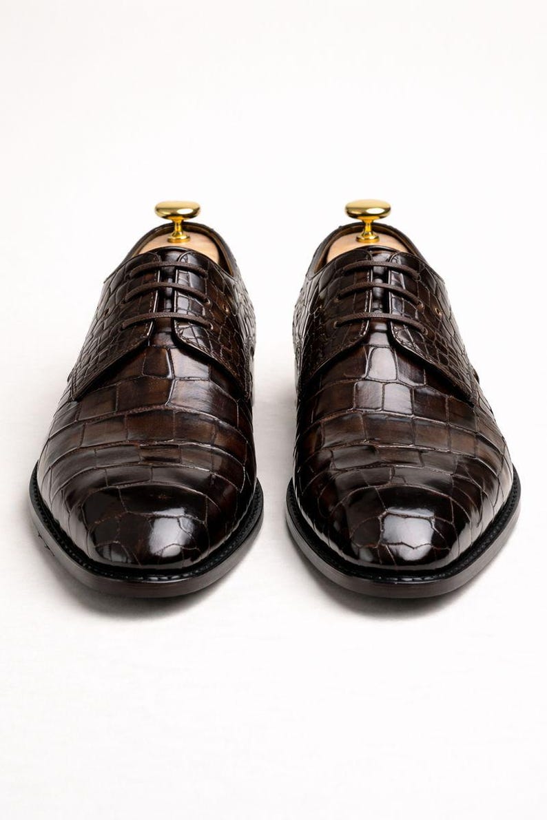 May include: A pair of brown leather dress shoes with a crocodile skin texture. The shoes are laced up and have a polished finish. Shoe trees are inserted inside the shoes. The background is white.
