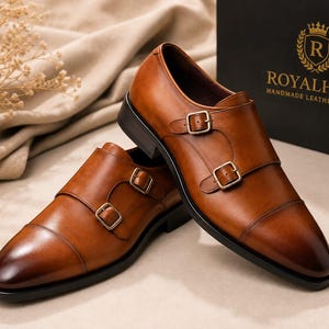May include: A pair of tan leather double monk strap shoes with a cap toe design and gold-toned buckles. The shoes are positioned near a black box featuring the gold text ROYALHIDE HANDMADE LEATHER SHOES and a circular logo with the letter R.