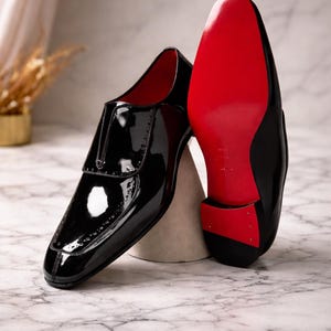 May include: A pair of black patent leather dress shoes with red soles. The shoes have a glossy finish and a classic design, suitable for formal occasions. The soles are a vibrant red.