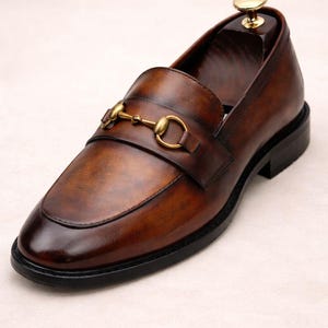 Puede incluir: Un solo mocas&iacute;n de cuero marr&oacute;n con un detalle de adorno met&aacute;lico dorado. El zapato tiene una puntera redondeada y una suela negra. El cuero tiene un acabado pulido y brillante. Se inserta una horma de zapato de madera dentro del zapato.
