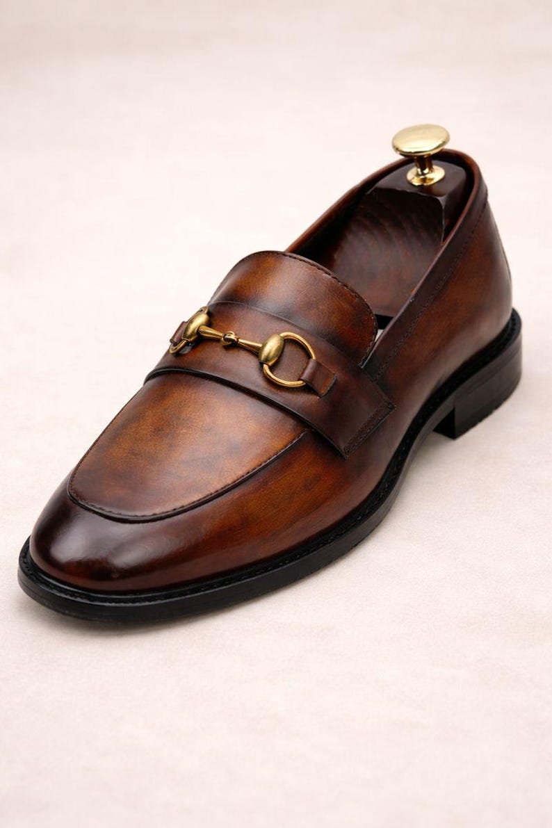 Puede incluir: Un solo mocas&iacute;n de cuero marr&oacute;n con un detalle de adorno met&aacute;lico dorado. El zapato tiene una puntera redondeada y una suela negra. El cuero tiene un acabado pulido y brillante. Se inserta una horma de zapato de madera dentro del zapato.