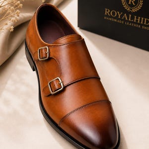 May include: A tan leather double monk strap shoe with a cap toe, featuring two brass-toned metal buckles on the side and a dark brown sole. The shoe is positioned near a black box with the gold text ROYALHID HANDMADE LEATHER SHOE.