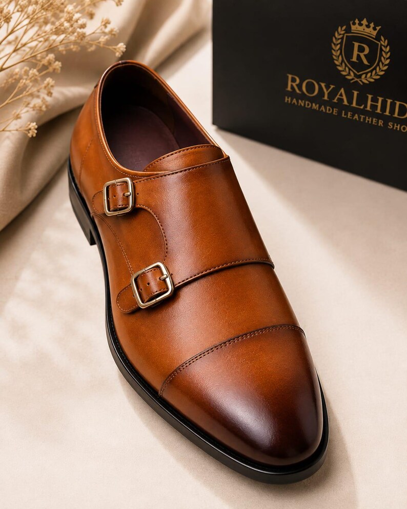 May include: A tan leather double monk strap shoe with a cap toe, featuring two brass-toned metal buckles on the side and a dark brown sole. The shoe is positioned near a black box with the gold text ROYALHID HANDMADE LEATHER SHOE.