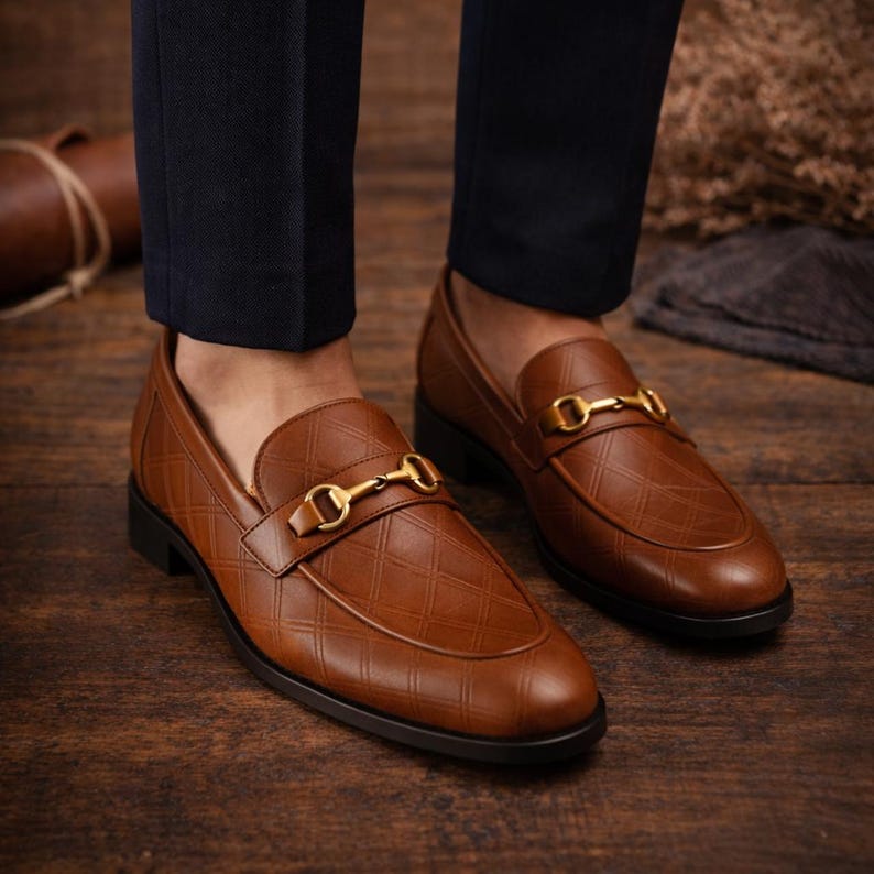 May include: Brown leather loafers with a diamond pattern and gold-colored metal accents. The shoes have a classic design with a rounded toe and a low heel. The loafers are shown on a wooden surface.