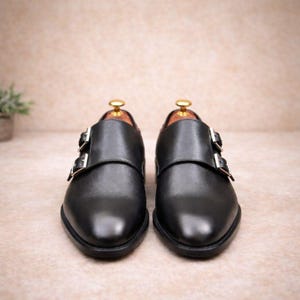 May include: A pair of black leather dress shoes with double monk straps. The shoes have a polished finish and silver-colored buckles. The shoes are displayed on a neutral background.