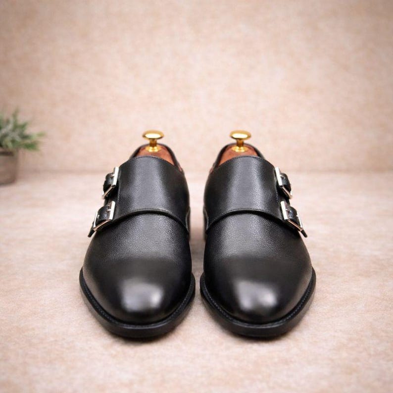 May include: A pair of black leather dress shoes with double monk straps. The shoes have a polished finish and silver-colored buckles. The shoes are displayed on a neutral background.
