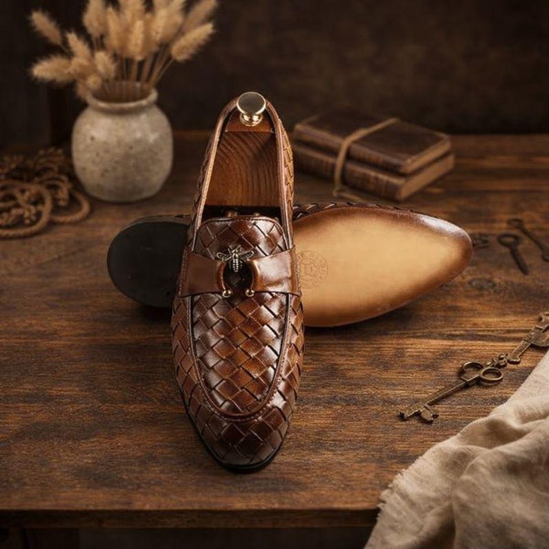 May include: Brown leather woven loafers with a decorative bee emblem. The shoes are displayed on a wooden surface with a blurred background. The loafers have a classic design and are suitable for formal or casual wear.