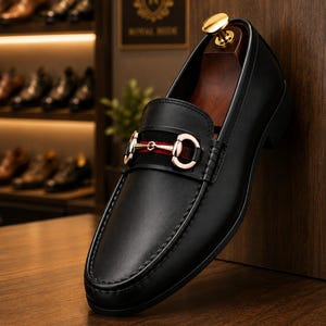 May include: A black leather loafer featuring a gold-toned metal horsebit detail across the vamp, set against a black suede strap with a red stripe. The shoe includes a wooden shoe tree insert with a gold-coloured knob at the heel.