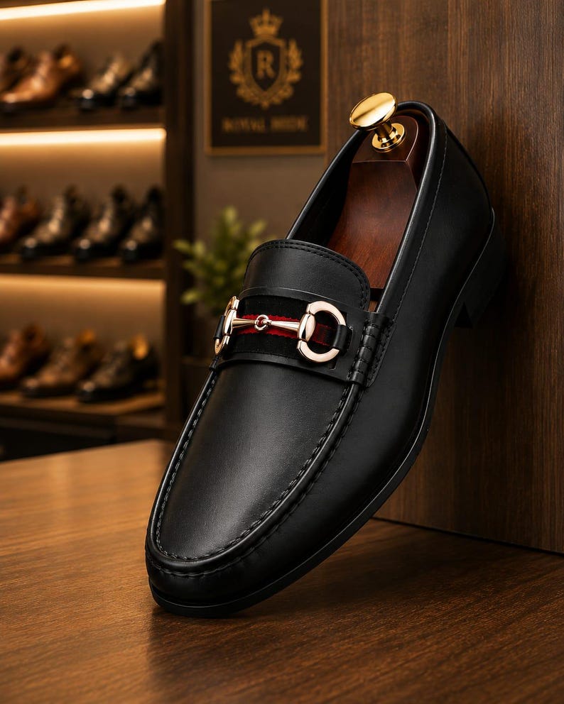 May include: A black leather loafer featuring a gold-toned metal horsebit detail across the vamp, set against a black suede strap with a red stripe. The shoe includes a wooden shoe tree insert with a gold-coloured knob at the heel.