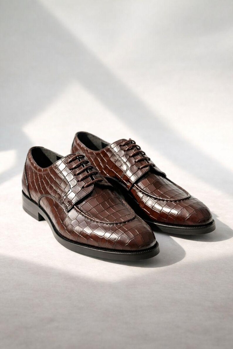 K&ouml;nnte beinhalten: Ein Paar braune Lederschuhe mit Krokodilleder-Textur. Die Schuhe haben ein klassisches Schn&uuml;rdesign und einen niedrigen Absatz. Die Schuhe werden auf einer wei&szlig;en Oberfl&auml;che pr&auml;sentiert.