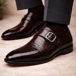 May include: Close-up of a pair of brown leather dress shoes with a crocodile texture. The shoes have a buckle closure and a red sole. The shoes are worn with dark gray dress pants and brown socks. The shoes are formal wear.