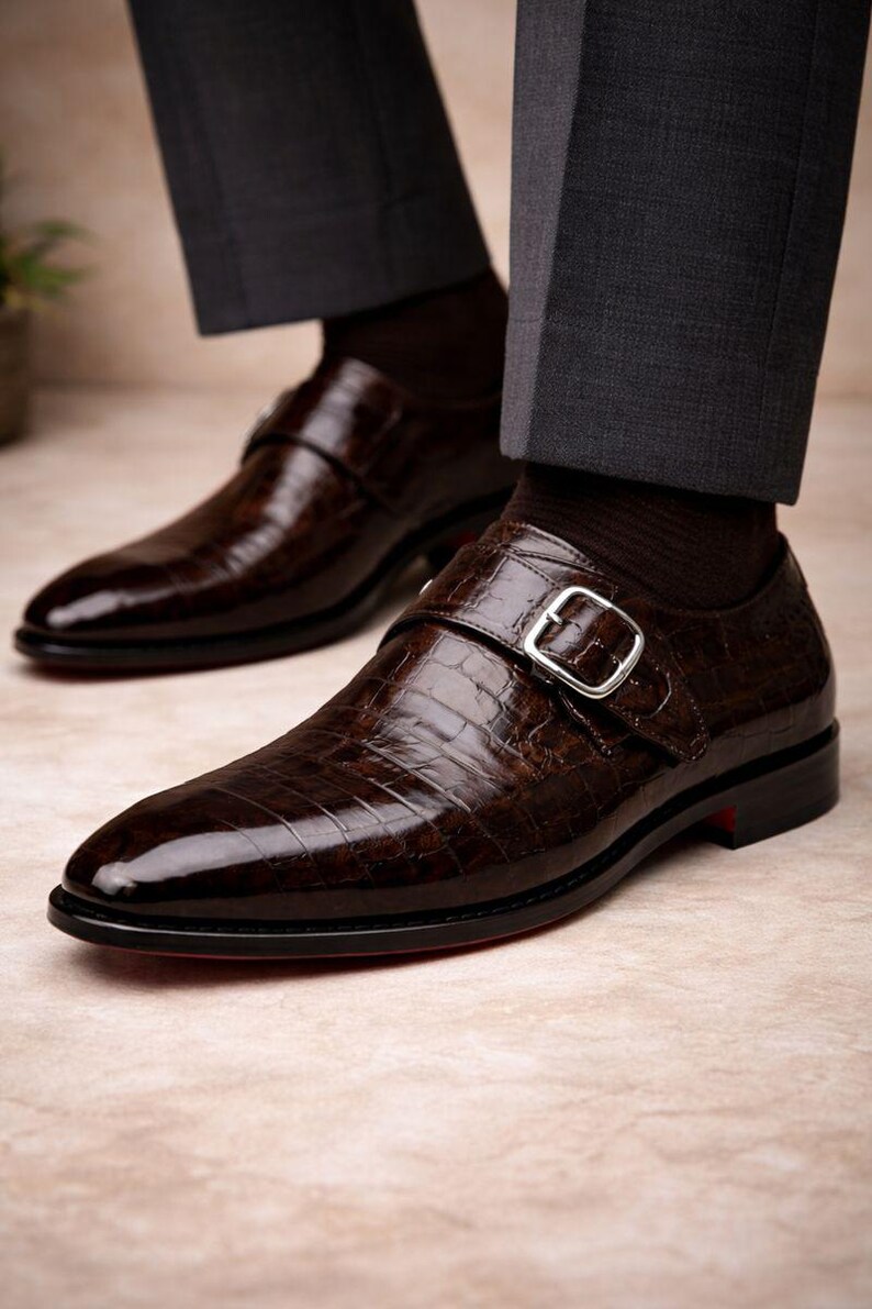 May include: Close-up of a pair of brown leather dress shoes with a crocodile texture. The shoes have a buckle closure and a red sole. The shoes are worn with dark gray dress pants and brown socks. The shoes are formal wear.