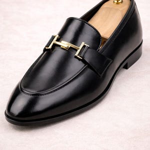 May include: Black leather loafer with a gold-colored buckle. The shoe has a rounded toe and a low heel. A wooden shoe tree is inside the shoe. The loafer is a classic style of footwear.