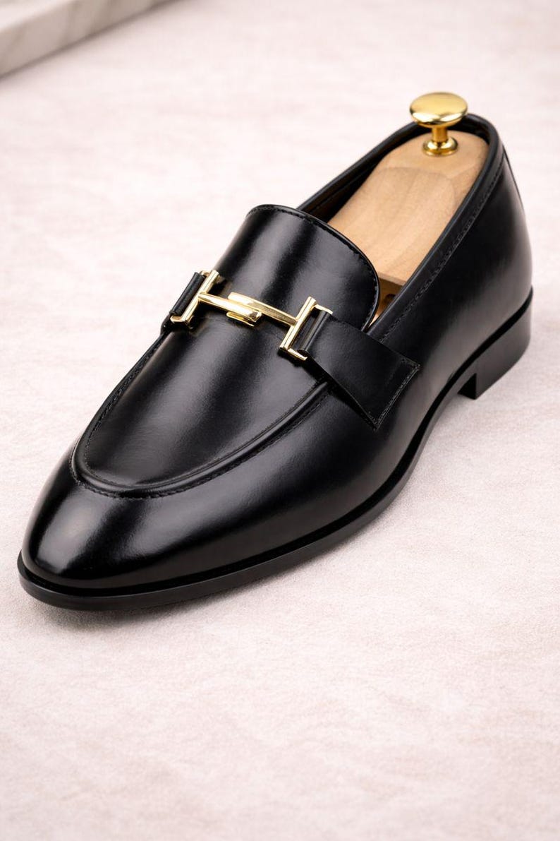 May include: Black leather loafer with a gold-colored buckle. The shoe has a rounded toe and a low heel. A wooden shoe tree is inside the shoe. The loafer is a classic style of footwear.