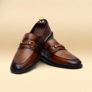 Puede incluir: Un par de mocasines de cuero marr&oacute;n con detalles met&aacute;licos dorados. Los zapatos tienen un dise&ntilde;o cl&aacute;sico con punta redondeada y una correa decorativa en la parte superior. El cuero tiene un acabado pulido y las suelas son negras.