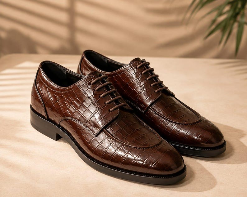 May include: A pair of dark brown dress shoes featuring a glossy crocodile-embossed texture, a rounded toe, and matching dark brown laces. The shoes have a low black heel and a structured silhouette with visible stitching along the vamp and sides.
