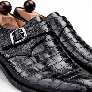 Op de afbeelding: Een paar zwarte leren herenschoenen met een krokodillenleerpatroon. De schoenen hebben een gespsluiting en worden gepresenteerd met houten schoenspanners. De schoenen hebben een glanzende afwerking en een klassiek ontwerp.