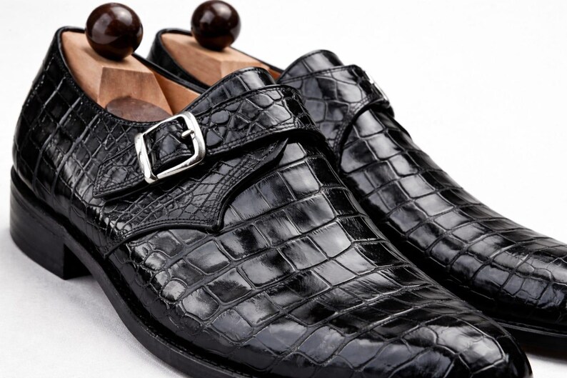 Op de afbeelding: Een paar zwarte leren herenschoenen met een krokodillenleerpatroon. De schoenen hebben een gespsluiting en worden gepresenteerd met houten schoenspanners. De schoenen hebben een glanzende afwerking en een klassiek ontwerp.