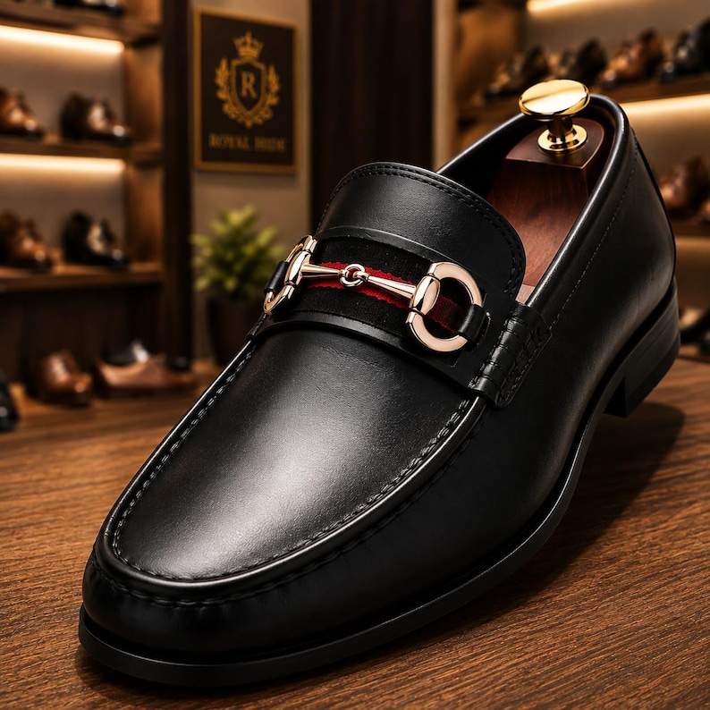 May include: A black leather loafer featuring a gold-toned metal horsebit detail across the vamp. The shoe includes a dark red fabric strap beneath the metal hardware and a wooden shoe tree inserted inside the heel area for structural support.