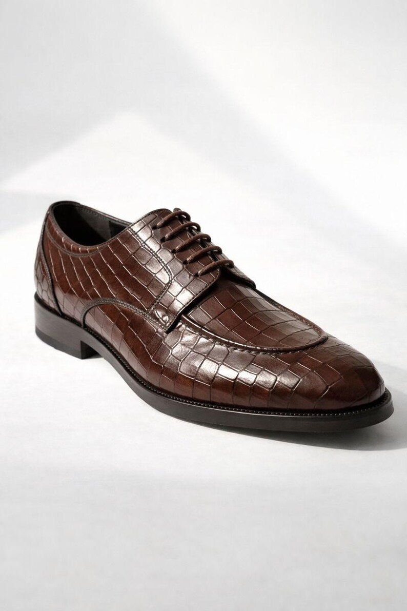 K&ouml;nnte beinhalten: Ein einzelner brauner Lederschuh mit einem strukturierten, krokodilartigen Muster. Der Schuh hat eine runde Spitze, einen Schn&uuml;rverschluss und eine dunkle Sohle. Der Schuh ist f&uuml;r formelle Anl&auml;sse konzipiert.