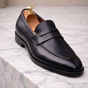 May include: A black leather loafer with a classic design. The shoe features a smooth upper, a strap across the vamp, and a low heel. A wooden shoe tree is inside the shoe. The shoe is displayed on a marble surface.