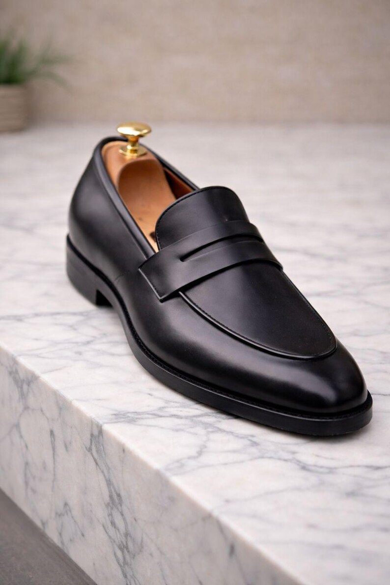 May include: A black leather loafer with a classic design. The shoe features a smooth upper, a strap across the vamp, and a low heel. A wooden shoe tree is inside the shoe. The shoe is displayed on a marble surface.