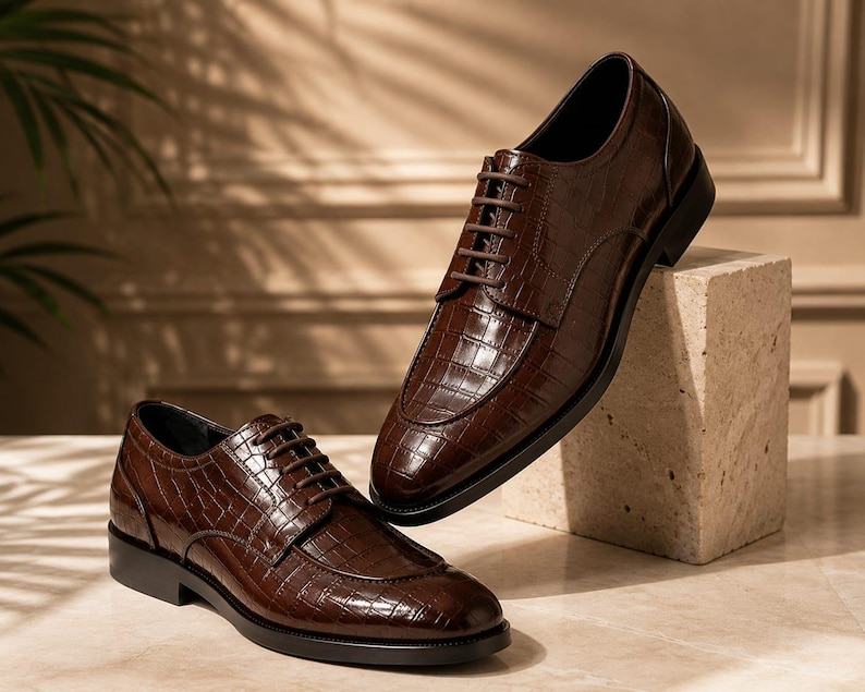 May include: A pair of dark brown lace-up dress shoes featuring a glossy crocodile-embossed leather texture. The shoes have a rounded toe, a low stacked heel, and matching dark brown laces, showcasing a structured silhouette and polished finish.
