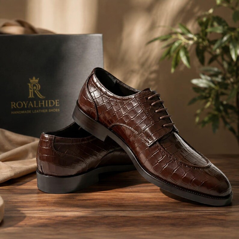 May include: A pair of dark brown lace-up shoes with a glossy crocodile-embossed texture. The shoes feature a rounded toe, a low heel, and matching brown laces. A black box with the gold text ROYALHIDE HANDMADE LEATHER SHOES is visible.