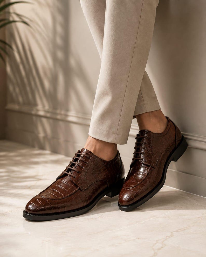 May include: A pair of dark brown leather oxford shoes featuring a glossy crocodile skin texture, a rounded toe, and matching dark brown laces. The shoes have a low black heel and a structured silhouette, providing a polished look for formal attire.