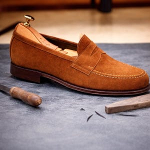 May include: A brown suede loafer with a leather sole and a stacked heel. The shoe features a classic penny loafer design with a stitched vamp and a leather lining. The shoe is approximately 11 inches long.