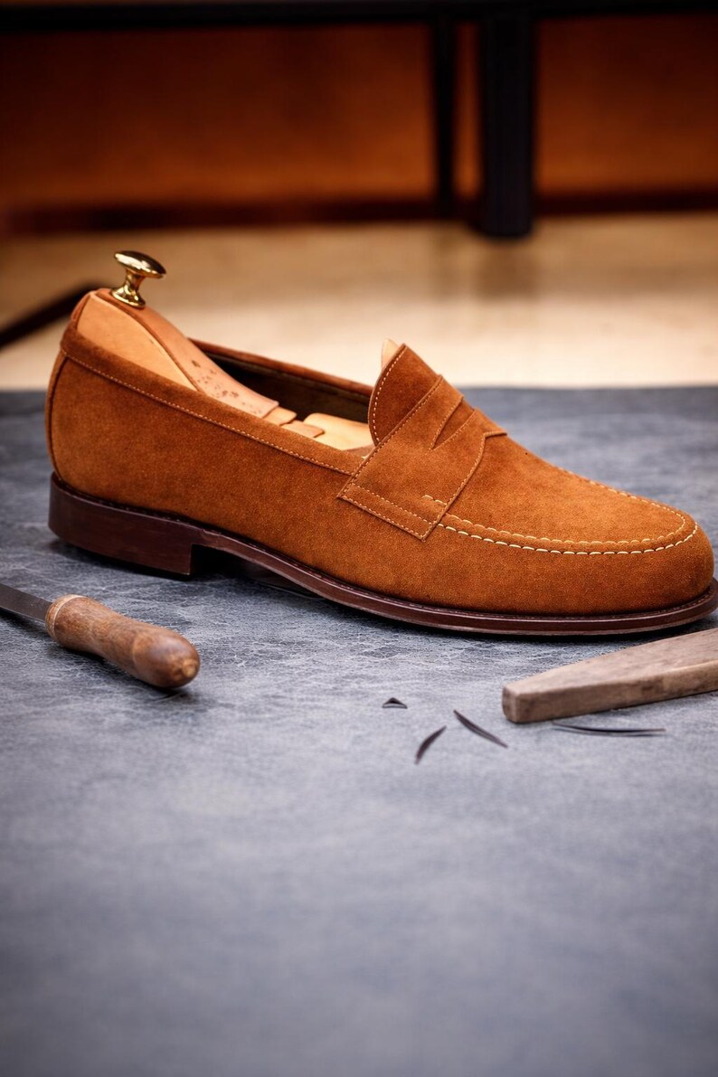 May include: A brown suede loafer with a leather sole and a stacked heel. The shoe features a classic penny loafer design with a stitched vamp and a leather lining. The shoe is approximately 11 inches long.