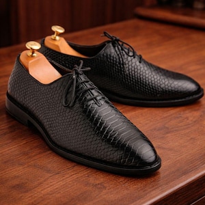 May include: A pair of black leather dress shoes with a textured, reptile-like pattern. The shoes have laces and are displayed on a wooden surface. Shoe trees are inserted inside the shoes to maintain their shape.