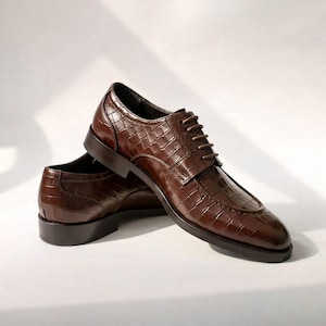 K&ouml;nnte beinhalten: Ein Paar braune Lederschuhe mit Krokodiltextur. Die Schuhe haben ein klassisches Schn&uuml;rdesign und eine dunkelbraune Sohle. Die Schuhe werden auf wei&szlig;em Hintergrund pr&auml;sentiert.