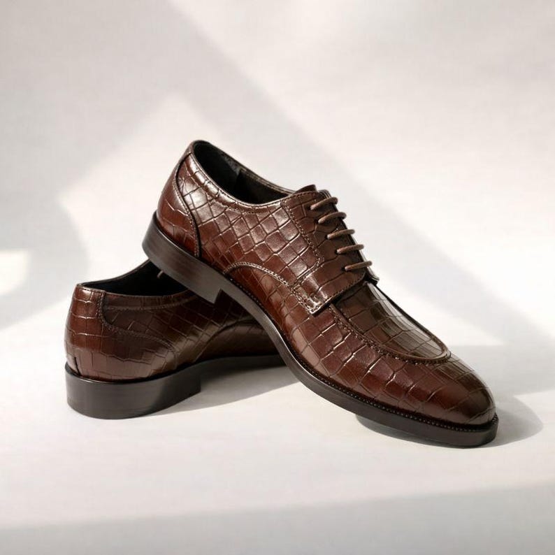 K&ouml;nnte beinhalten: Ein Paar braune Lederschuhe mit Krokodiltextur. Die Schuhe haben ein klassisches Schn&uuml;rdesign und eine dunkelbraune Sohle. Die Schuhe werden auf wei&szlig;em Hintergrund pr&auml;sentiert.