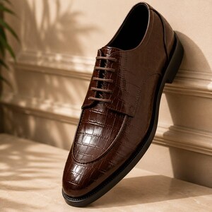 May include: A dark brown dress shoe with a glossy finish and a textured crocodile skin pattern. The shoe features a rounded toe, a lace-up closure with thin brown laces, and a solid black sole. The design includes a stitched apron front detail.