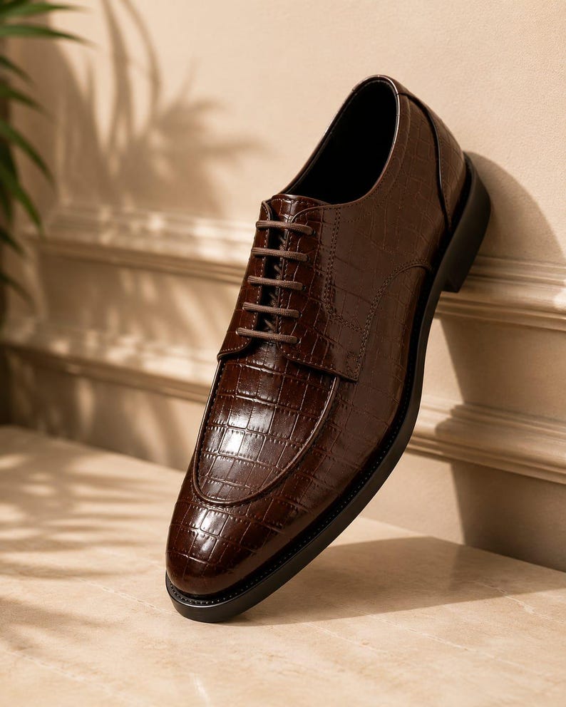 May include: A dark brown dress shoe with a glossy finish and a textured crocodile skin pattern. The shoe features a rounded toe, a lace-up closure with thin brown laces, and a solid black sole. The design includes a stitched apron front detail.