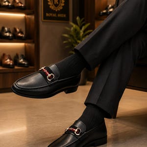 May include: Black leather loafers featuring a gold-tone metal horsebit detail across the vamp, accented by a horizontal red and green striped fabric band. The shoes are worn with black ribbed socks and paired with black trousers.