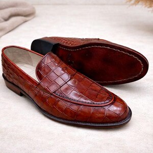 May include: A pair of brown leather loafers with a crocodile pattern. One shoe is upright, showcasing the textured upper and red trim, while the other is inverted, revealing the sole. The shoes are on a light-colored surface.