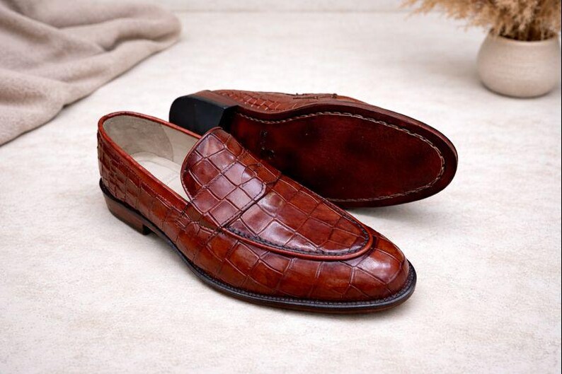 May include: A pair of brown leather loafers with a crocodile pattern. One shoe is upright, showcasing the textured upper and red trim, while the other is inverted, revealing the sole. The shoes are on a light-colored surface.