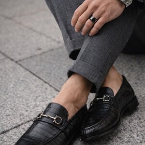 May include: Black leather loafers with a crocodile texture and gold-colored metal accents. The shoes are paired with gray trousers. The loafers have a classic design, suitable for formal or semi-formal occasions.