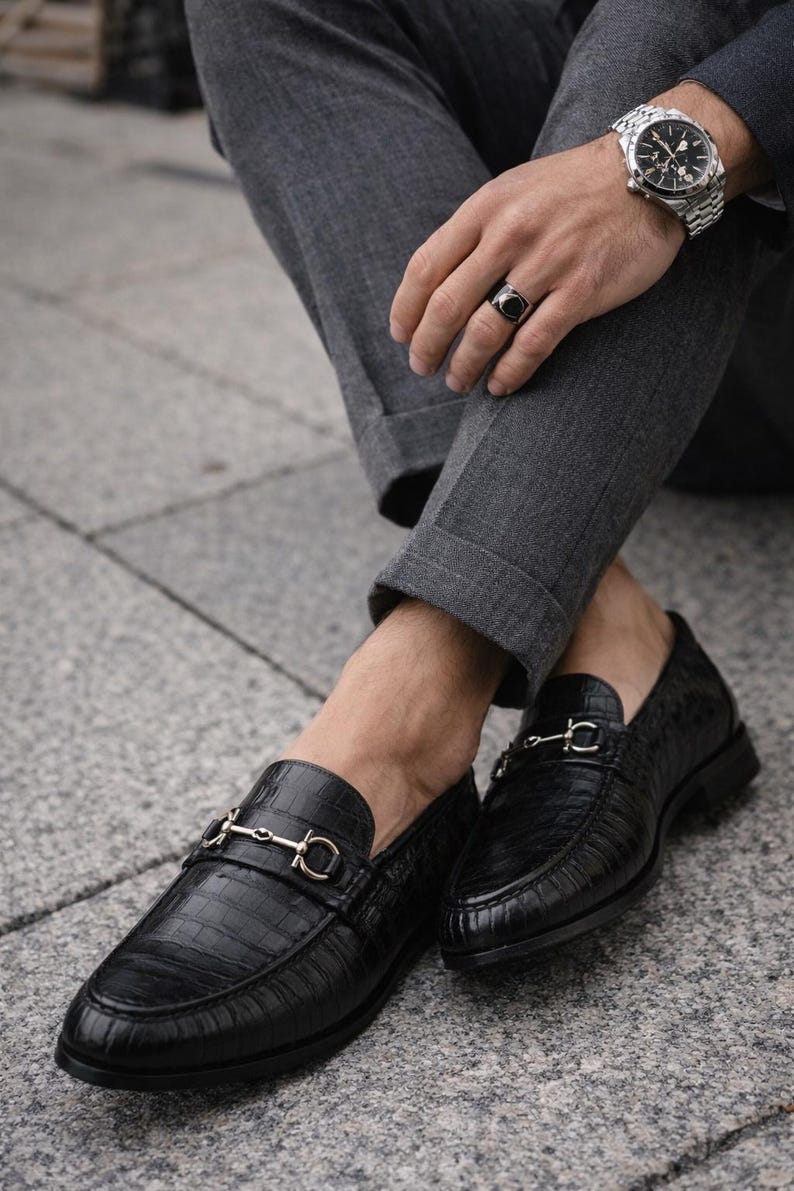 May include: Black leather loafers with a crocodile texture and gold-colored metal accents. The shoes are paired with gray trousers. The loafers have a classic design, suitable for formal or semi-formal occasions.